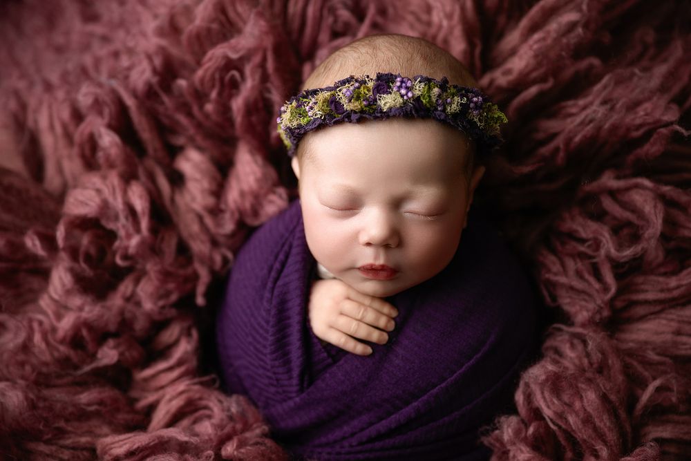 Sleeping newborn wrapped in purple fabric with ornate headband on burgundy textured backdrop.
