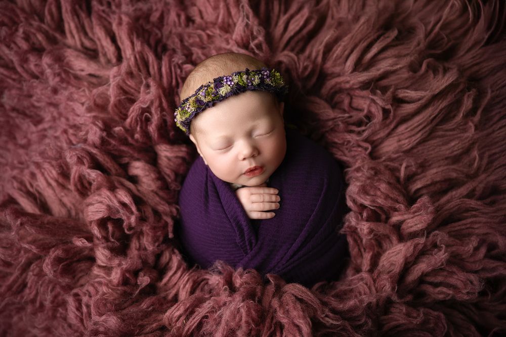 Newborn swaddled in deep purple wrap with floral crown on burgundy fur background.
