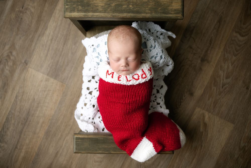 Newborn wearing red and white Christmas stocking outfit lying on wooden surface with lace accent.