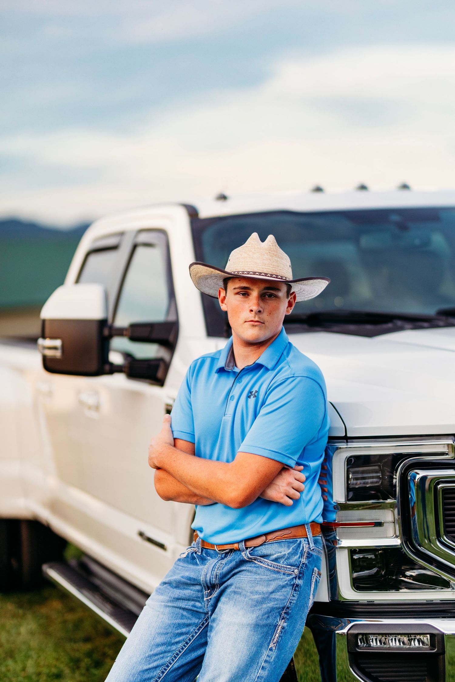 Young man in cowboy hat and blue polo leaning on white truck with arms crossed.