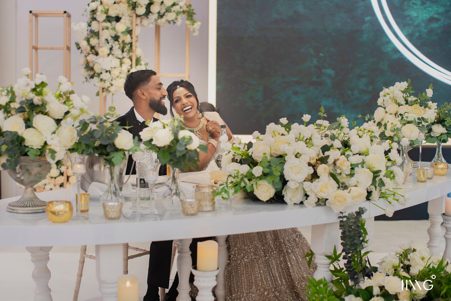 A couple shares a romantic moment at their wedding reception decorated with lush white floral arrangements and modern decor.