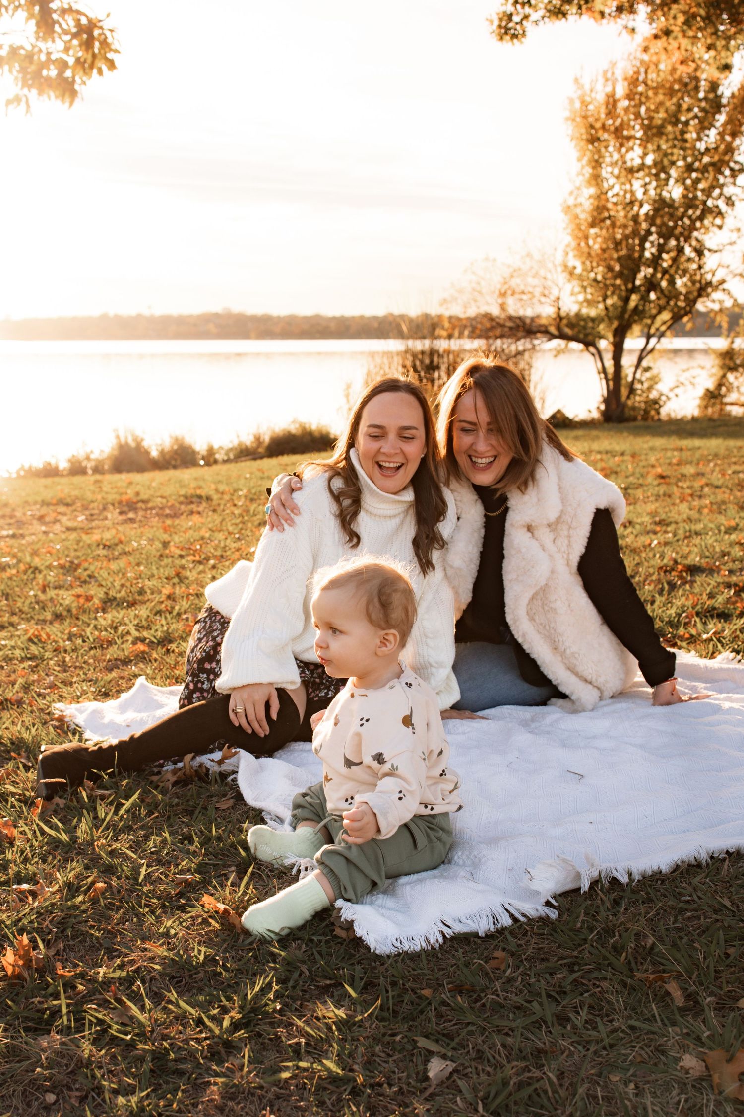 Family sitting on a white blanket by a lake during golden hour sunset.