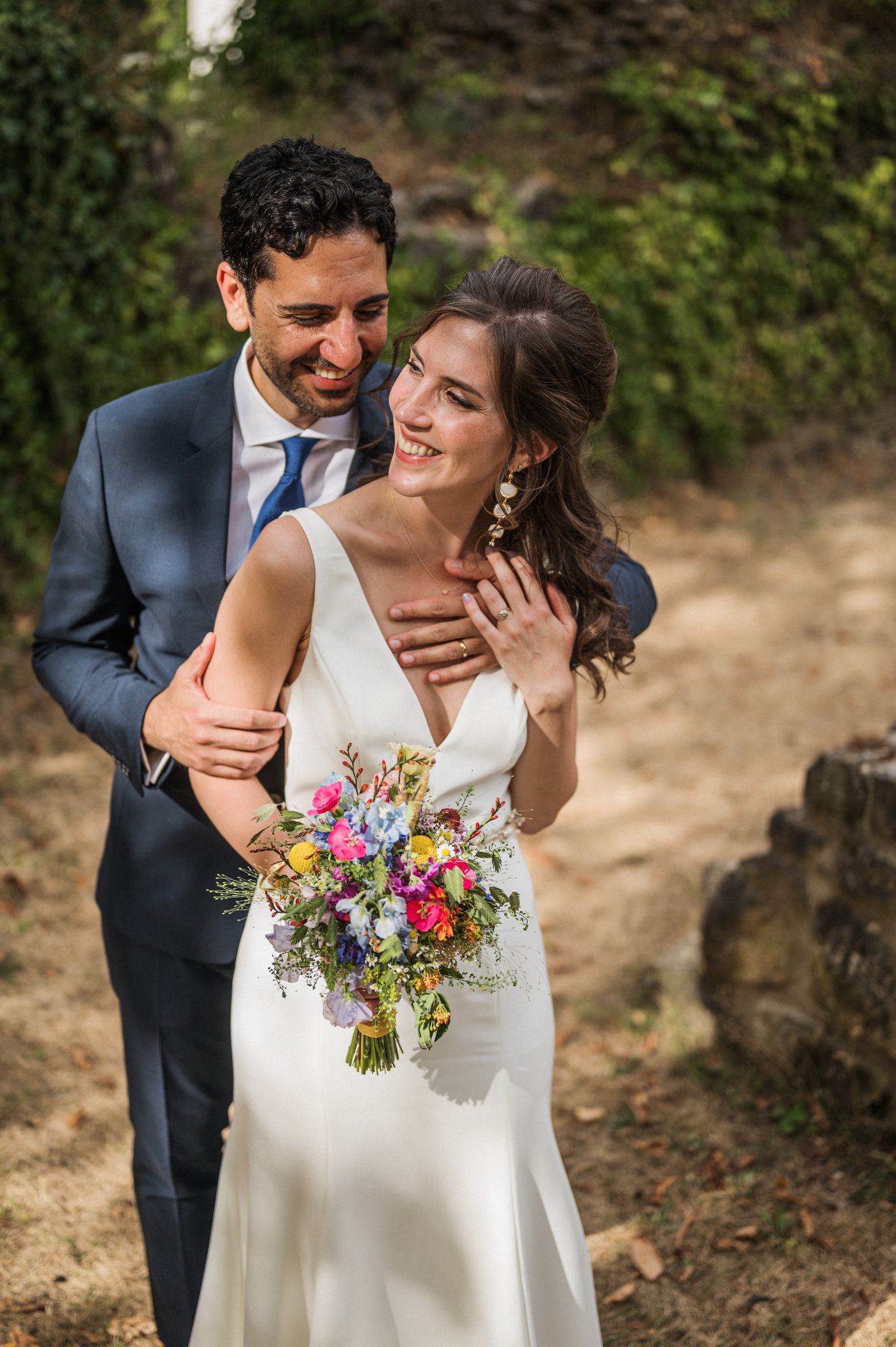 A joyful wedding couple embraces outdoors while the bride holds a vibrant wildflower bouquet in her white dress.
