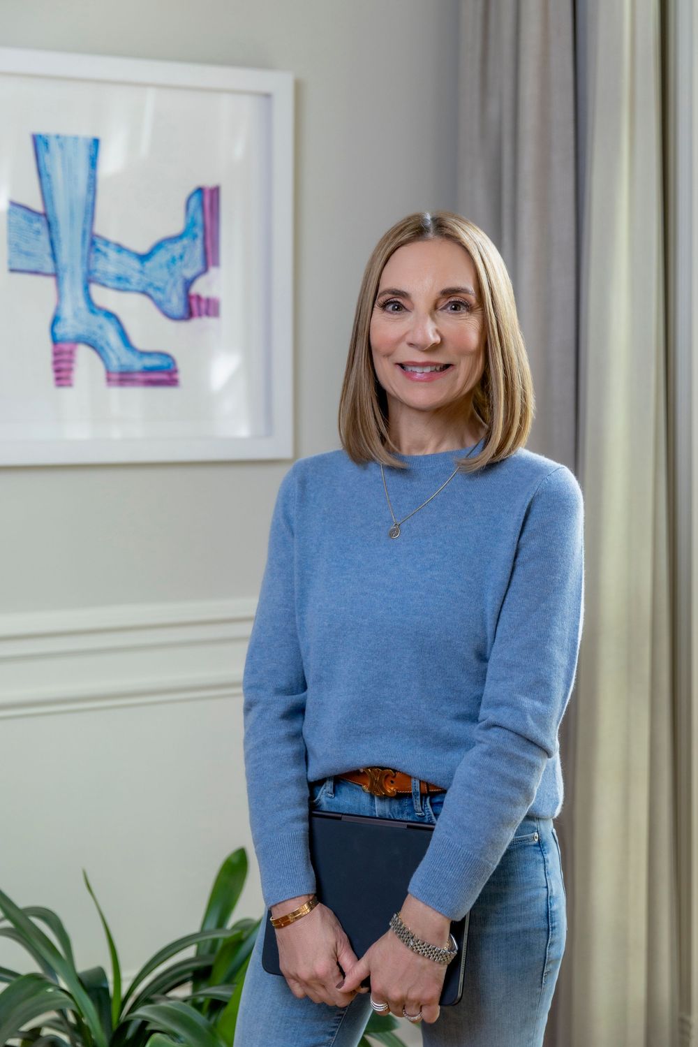 Person in blue sweater and dark pants stands in office setting with framed artwork and plants in background.