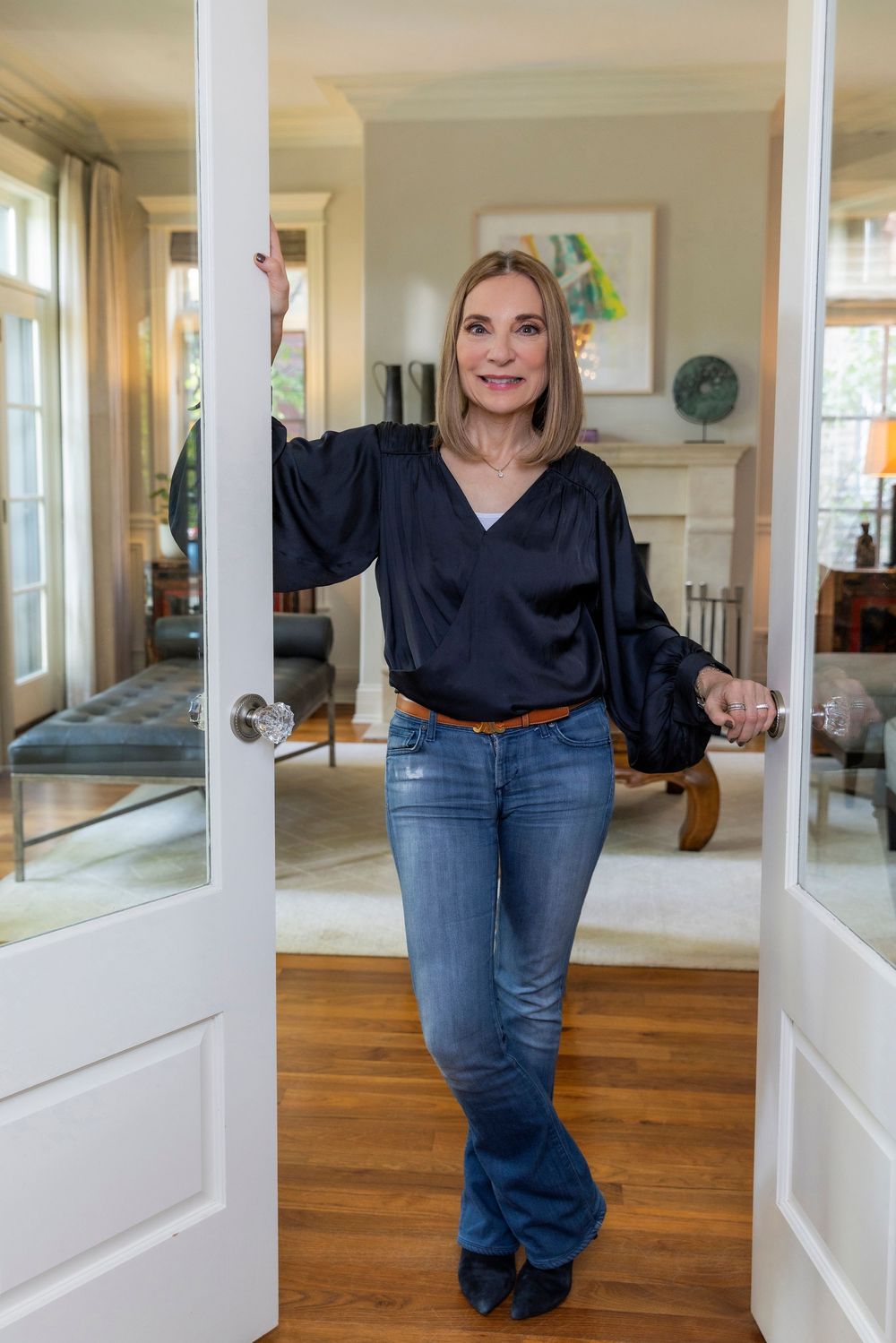 Woman in casual outfit with black blouse and blue jeans stands in doorway of bright living room interior.