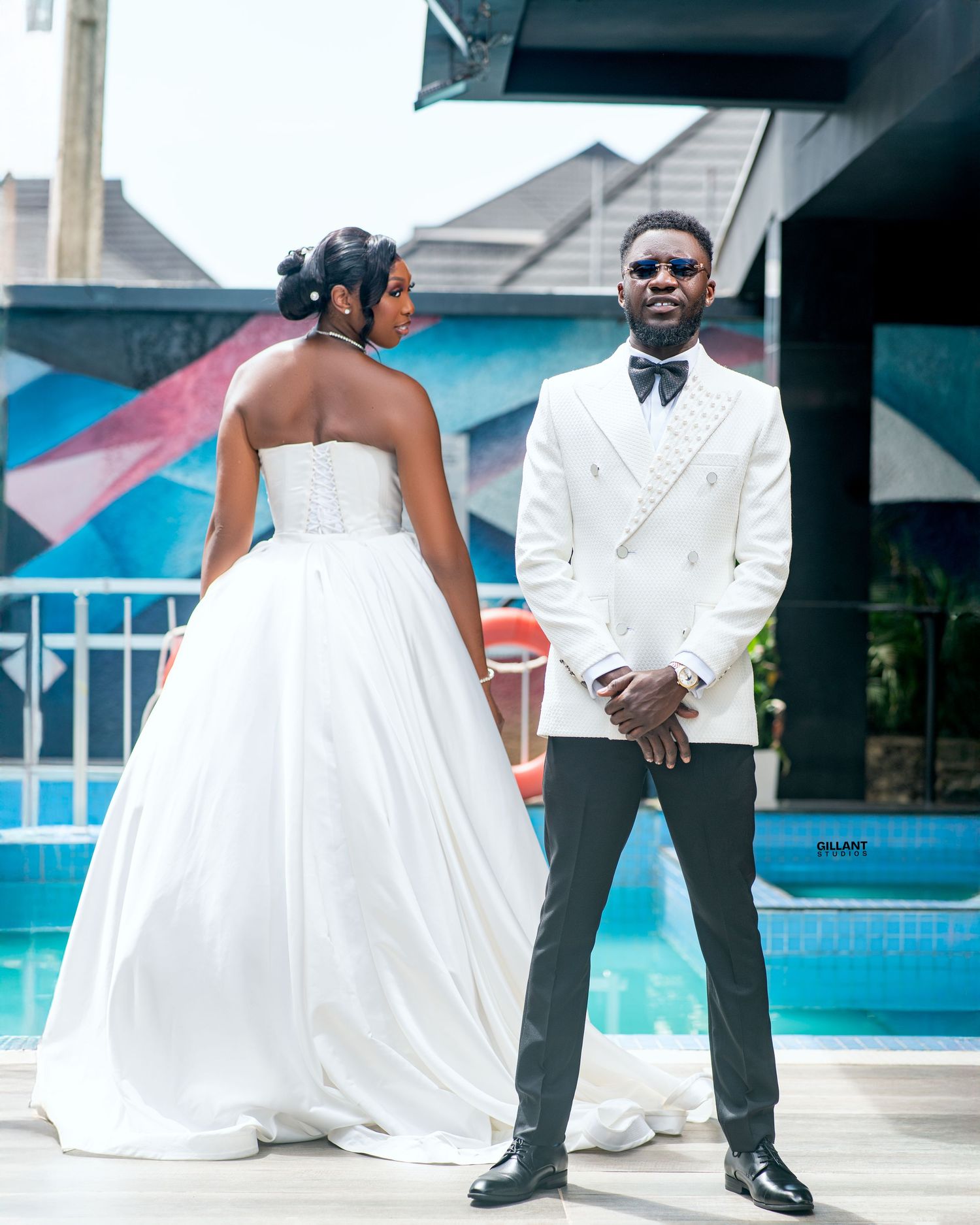 Elegant couple in formal white wedding attire pose by poolside setting.
