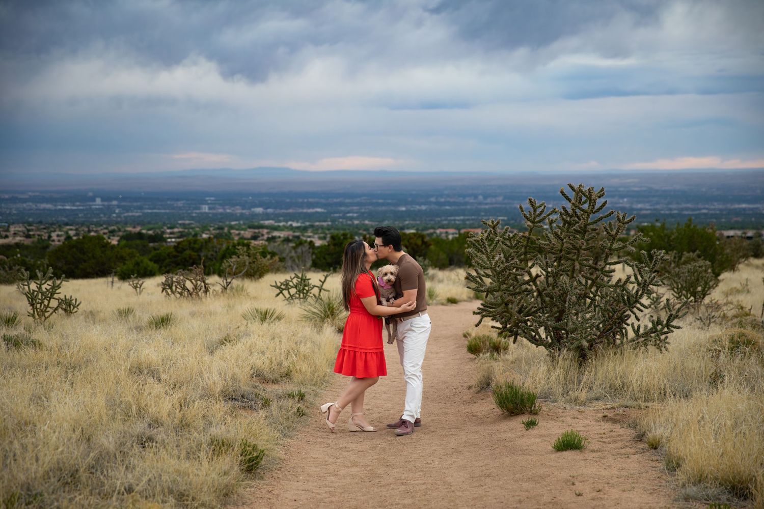 Couple sharing a romantic kiss on a desert trail with sweeping valley views and dramatic cloudy skies in New Mexico.