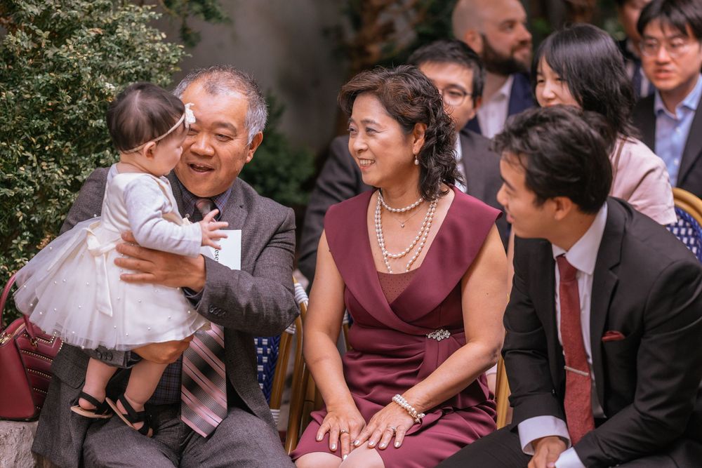 Asian family smiling at a baby during an outdoor wedding ceremony, grandparents holding infant in white dress.