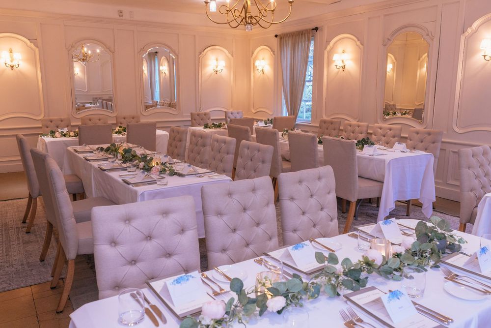 Elegant wedding reception room with tufted chairs, white tablecloths, greenery garlands, and gold chandeliers in a cream-colored venue.