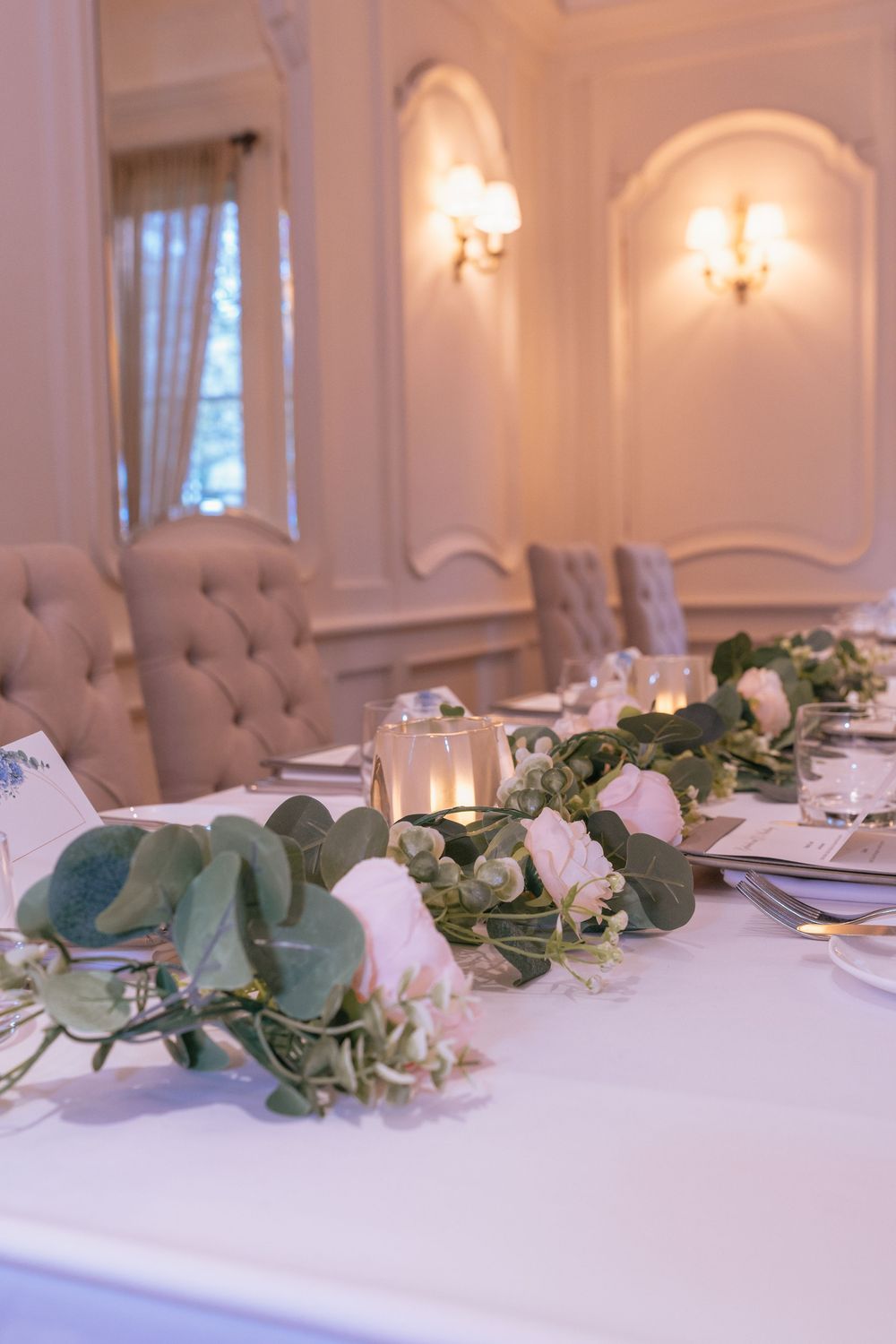 Elegant wedding reception table with eucalyptus garland, white roses, and glowing candles in a warmly lit ballroom.