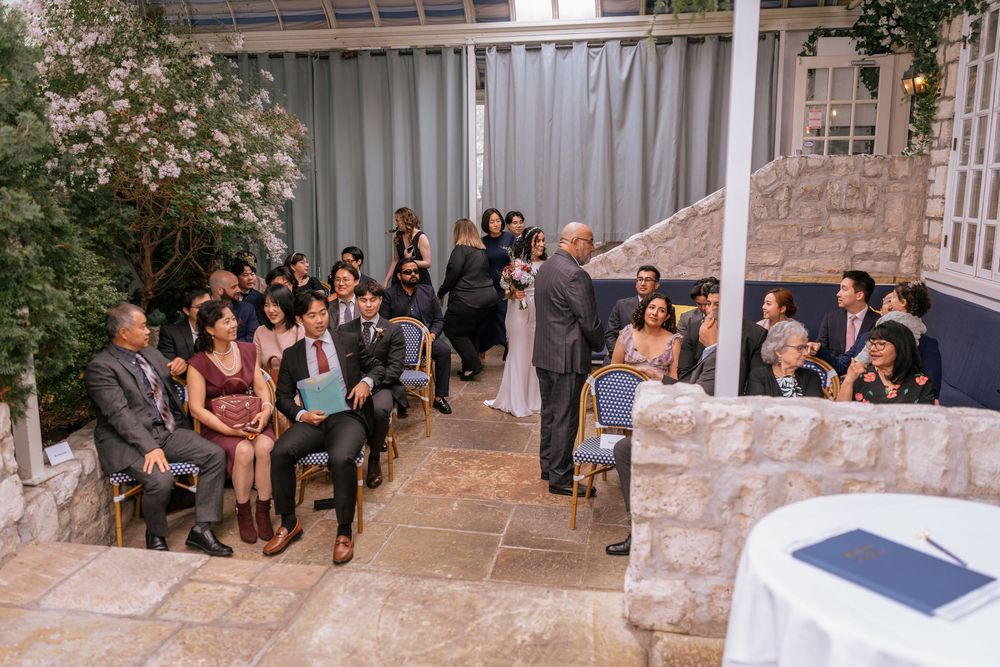Elegant indoor wedding ceremony with guests seated in chairs at a stone-walled venue with lush greenery.