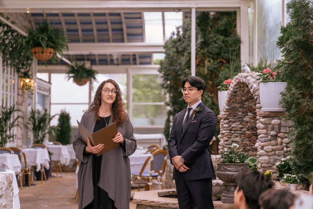 A woman officiant holds a folder while standing beside a groom in a suit at an indoor garden wedding venue.