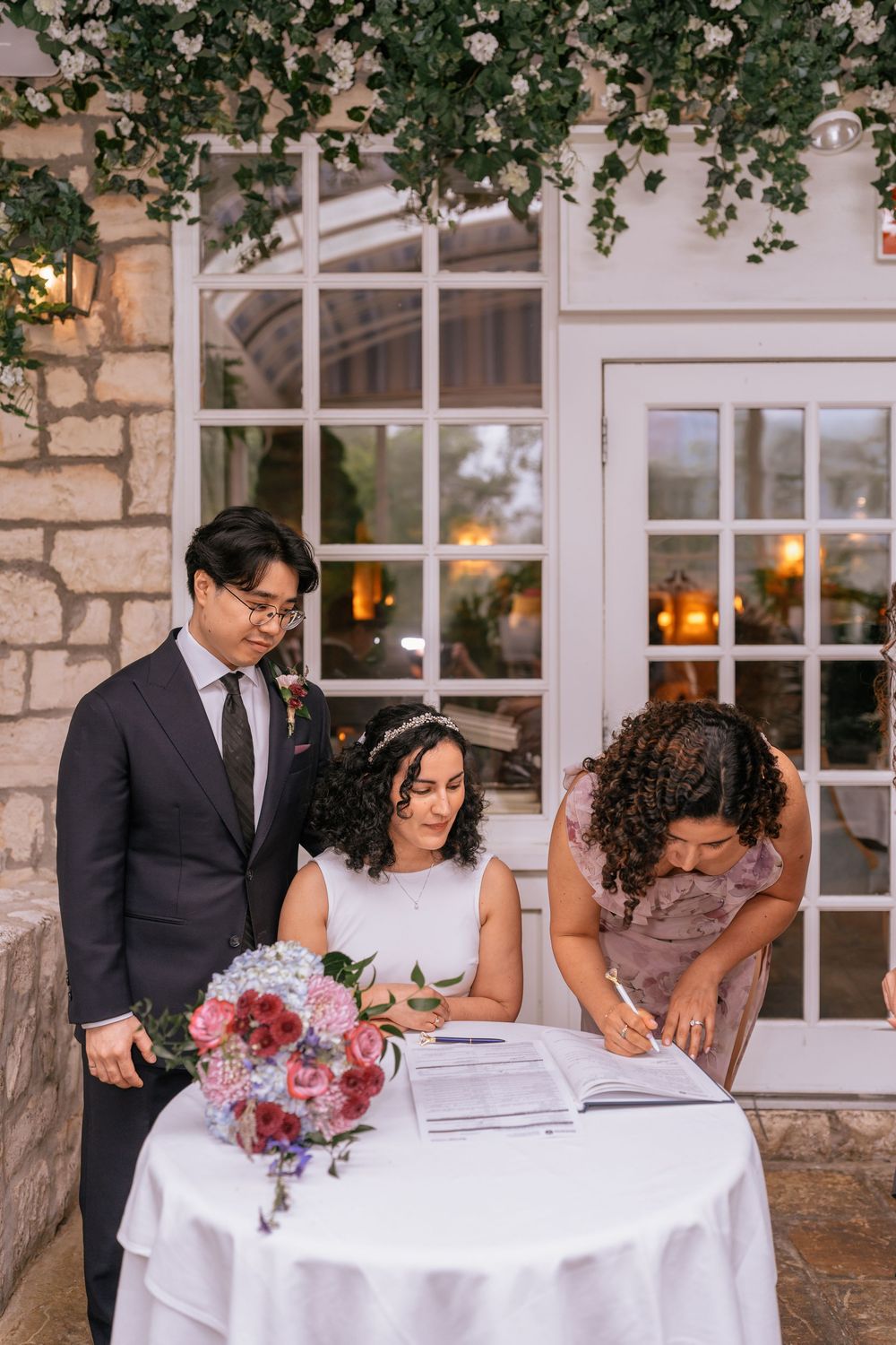 Bride signs wedding registry at white table with groom and witness, surrounded by floral decor and French doors.