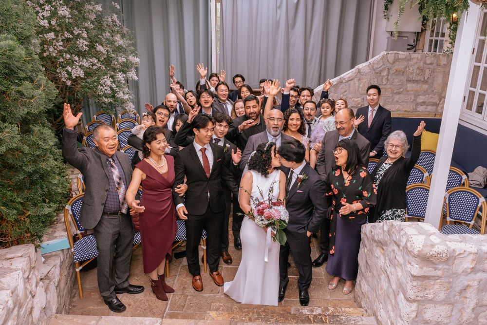 A joyful wedding group photo with guests cheering as the bride and groom share a kiss on stone steps.