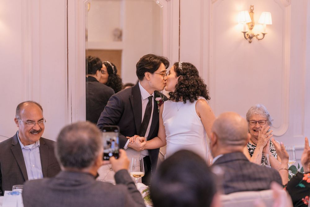Newlyweds share a kiss at their wedding reception as smiling guests applaud and celebrate around them.