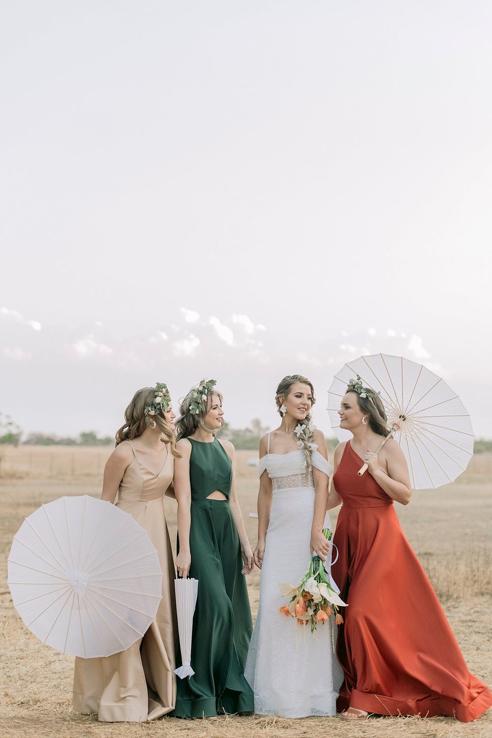 Bride with three bridesmaids in colorful gowns holding white parasols in an open golden field.