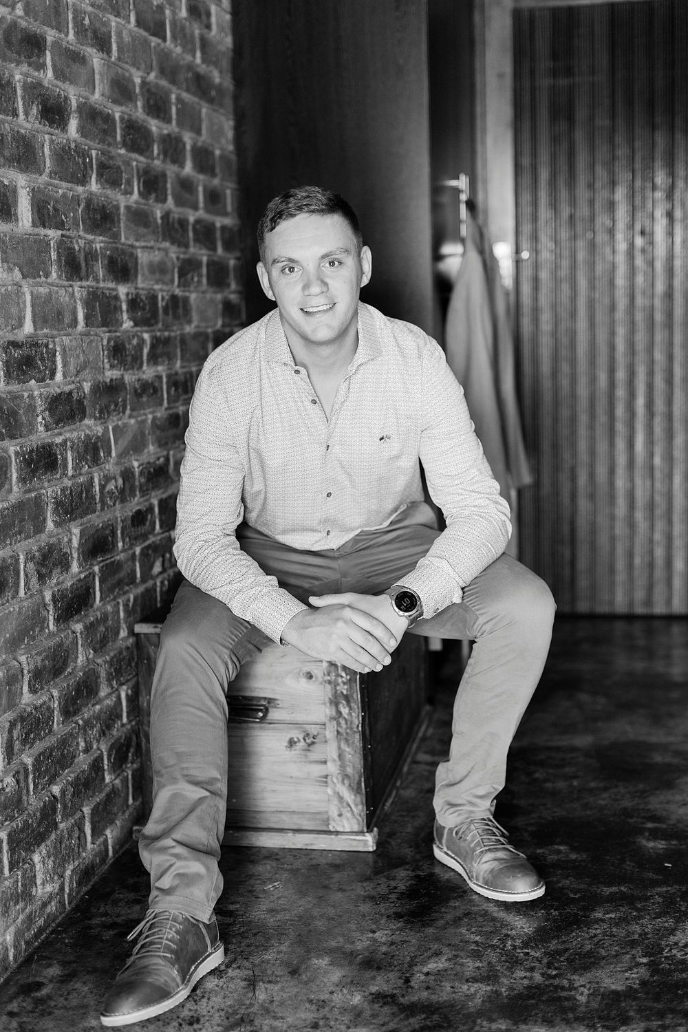 Young man in casual shirt sitting on wooden crate against brick wall in black and white portrait.