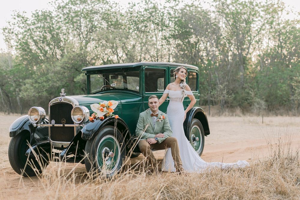 Bride and groom posing beside a vintage green classic car decorated with flowers in a golden field.