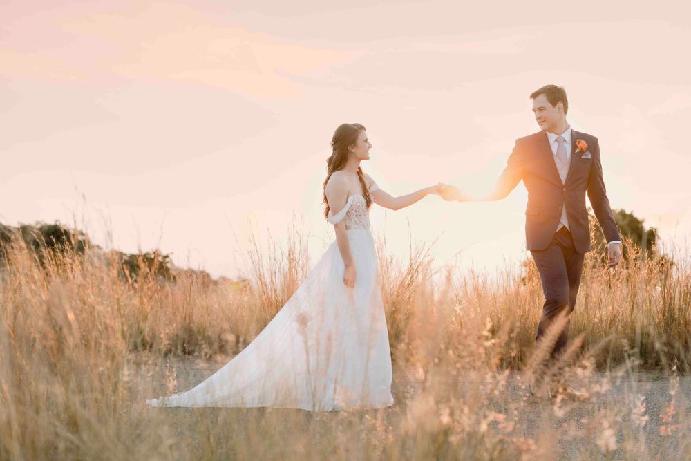 Bride and groom holding hands walking through golden wheat field during a stunning sunset.