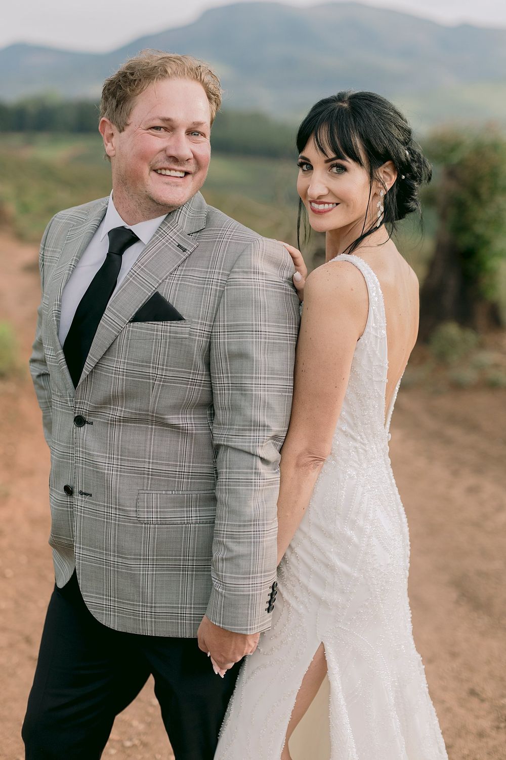 Smiling bride and groom posing on a dirt road with mountain vineyard backdrop.