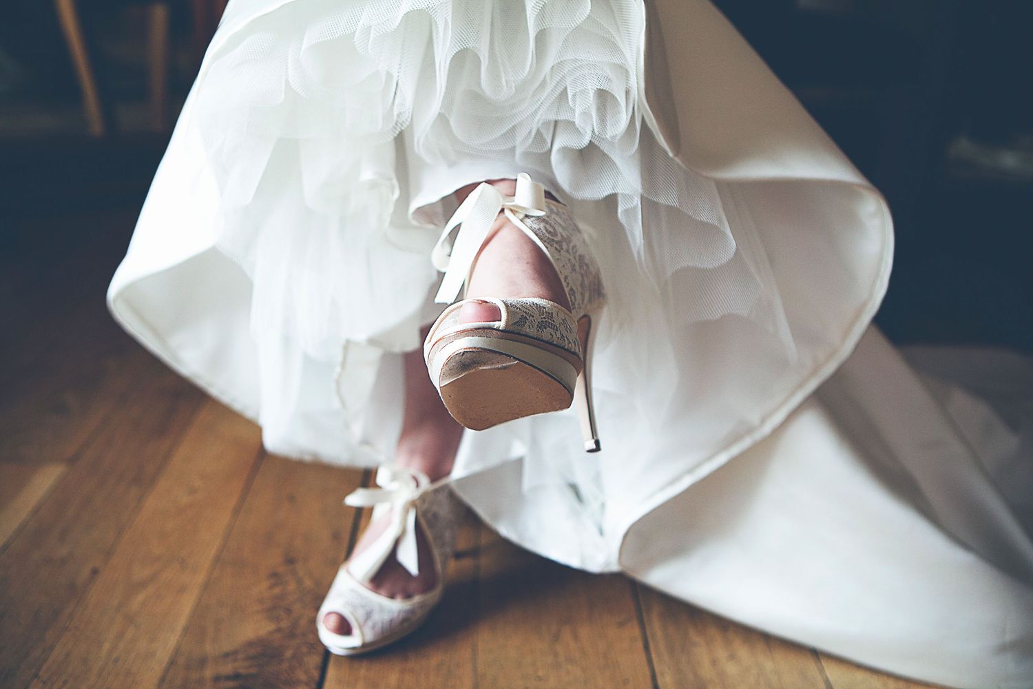 Bride wearing elegant white peep-toe heels with bow detail beneath a flowing ruffled wedding gown on wooden floor.
