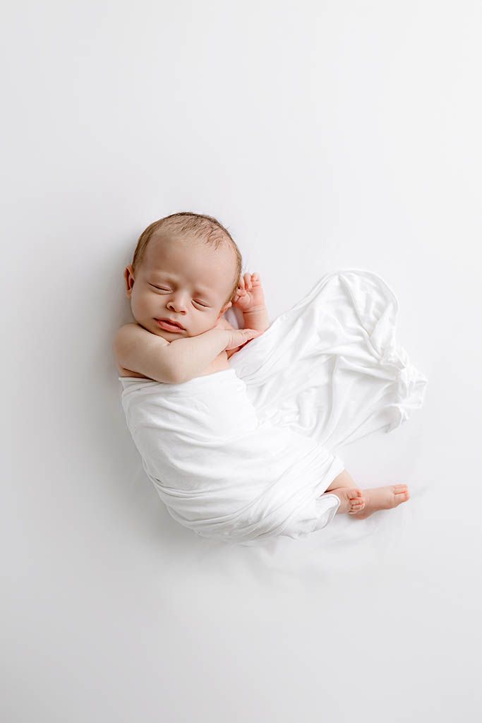 Peaceful newborn baby sleeping wrapped in white cloth on white background, curled in fetal position.