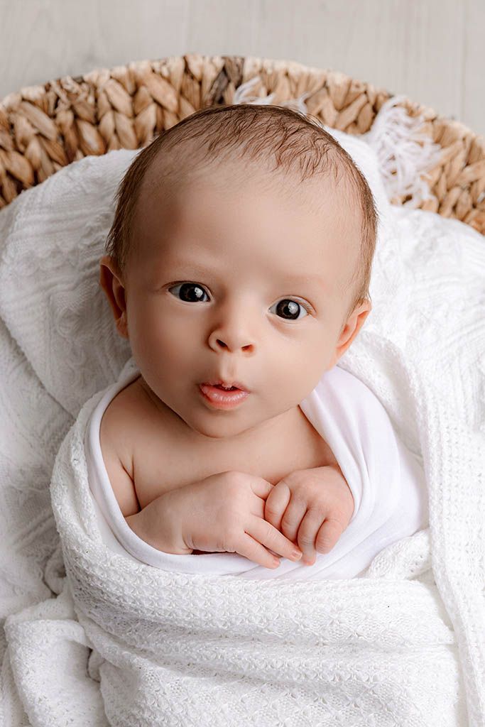 Newborn baby wrapped in white towel lying in wicker basket with alert dark eyes and tiny hands visible.