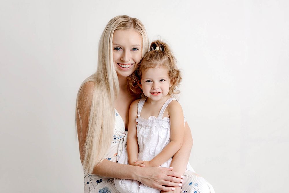 Smiling blonde woman hugging a curly-haired little girl in white dresses against a light background.