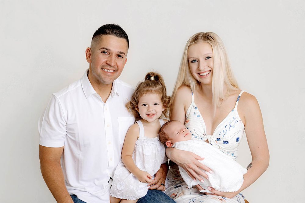 Happy family of four posing for a newborn portrait, parents with toddler daughter and swaddled baby against white background.