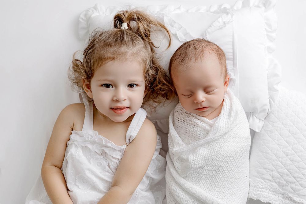 Two young siblings, a curly-haired toddler girl and a sleeping newborn wrapped in white, lying together on a white bed.