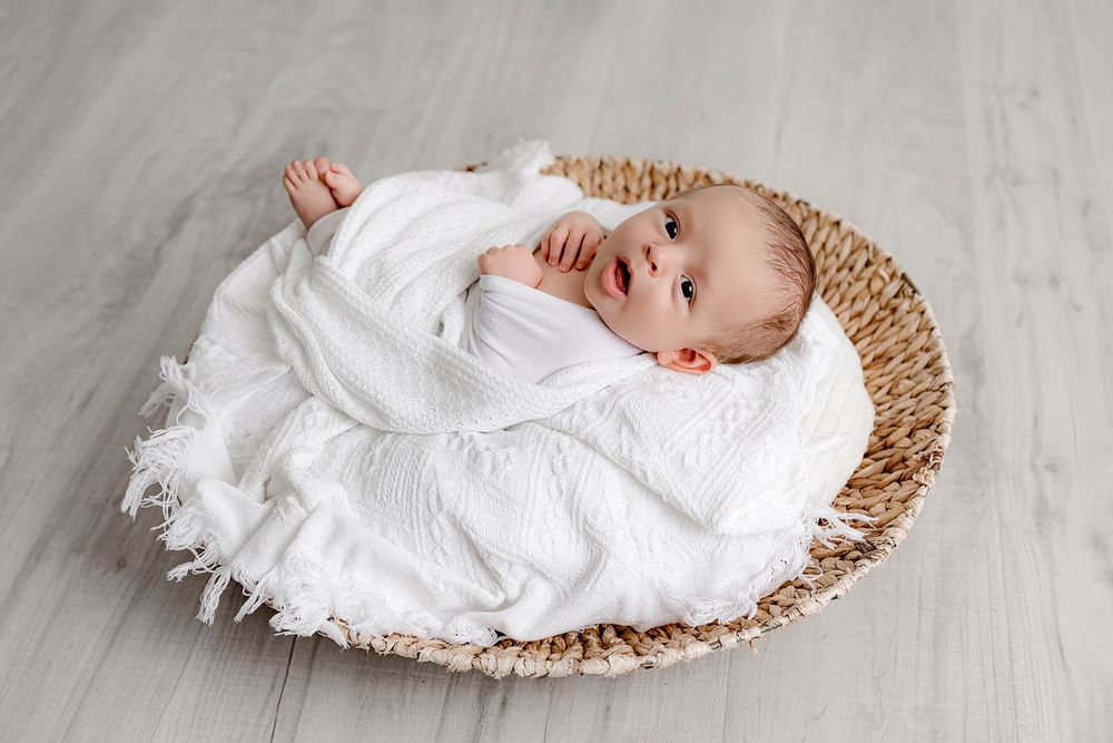 Adorable newborn baby wrapped in white muslin cloth lying in a woven basket on a light wood floor.