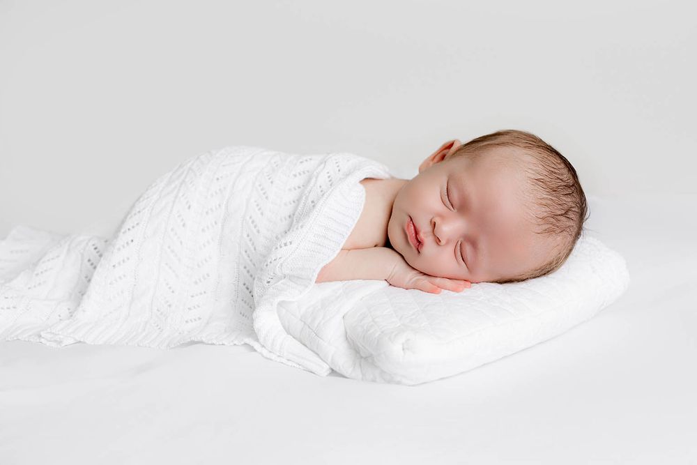 Newborn baby sleeping peacefully on a white pillow, swaddled in a soft white blanket against a light background.