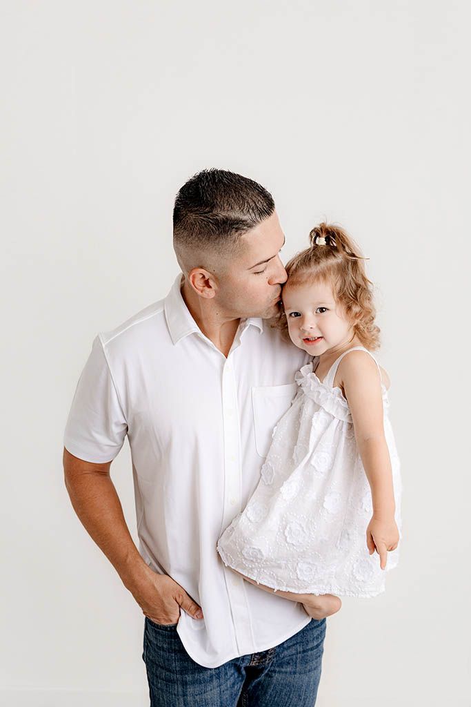 Father in white shirt kissing young daughter in white dress while holding her against a white background.