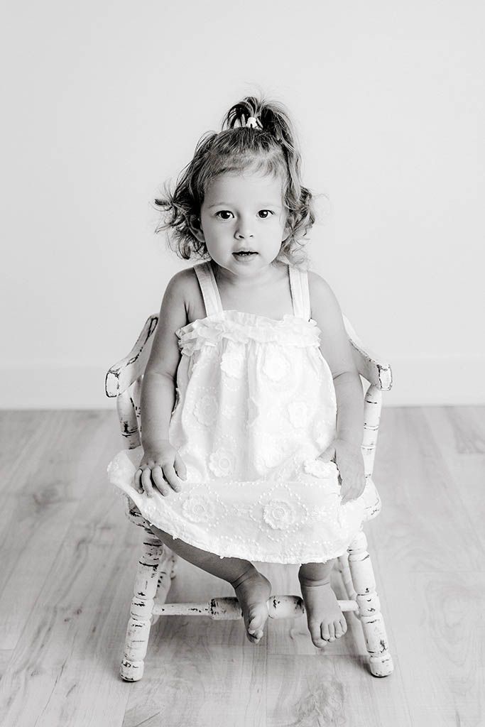 Black and white portrait of a curly-haired toddler girl in white dress sitting in a small chair.