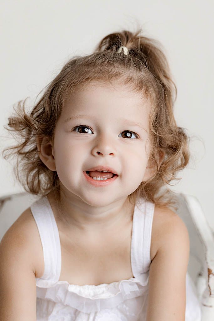 Cute toddler girl with curly hair in a white dress smiling during a studio portrait session.