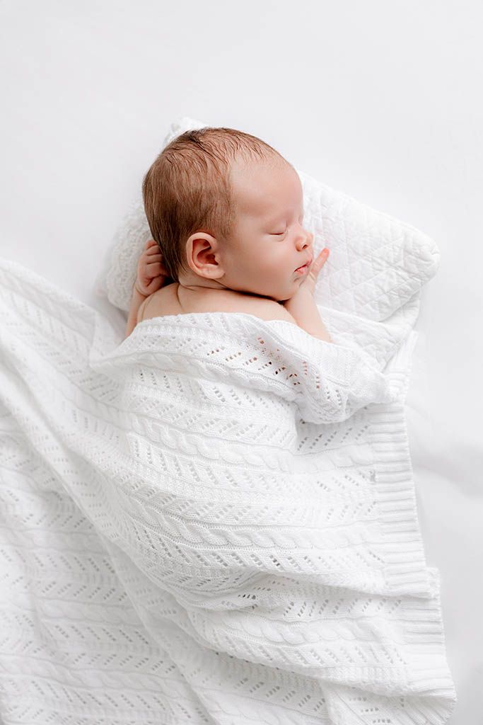 Peaceful newborn baby sleeping wrapped in white knit blanket on white surface.