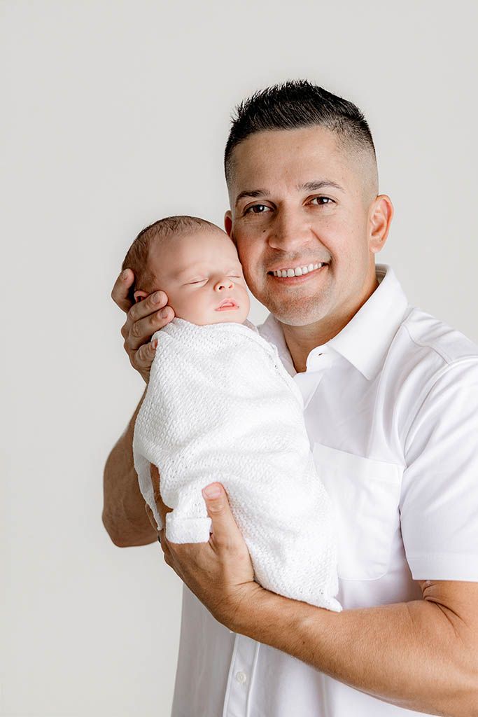 Smiling father in white shirt holding swaddled newborn baby against light background.