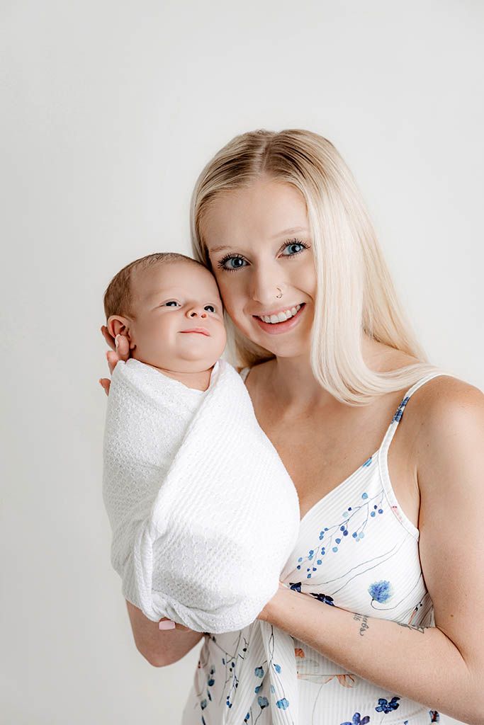 Smiling blonde mother holding her swaddled newborn baby in a white floral dress against a light background.