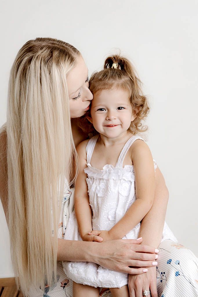 Blonde mother kissing cheek of smiling toddler girl in white dress.
