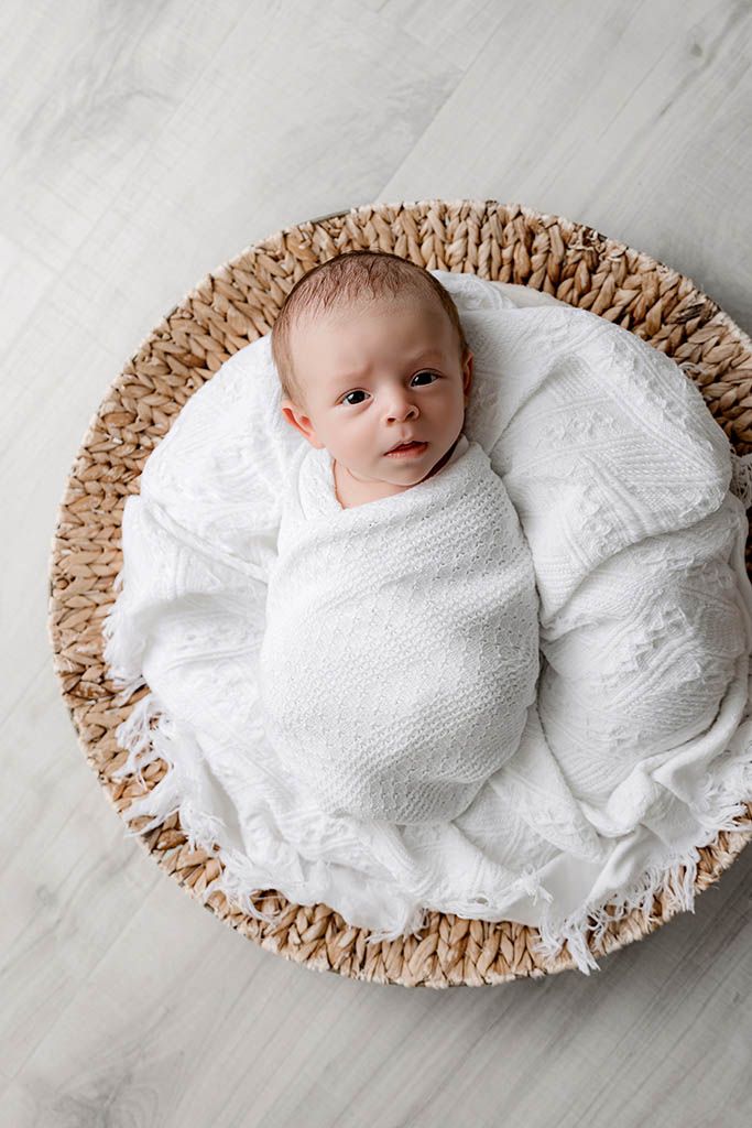 Awake newborn baby swaddled in white wrap lying in woven basket.