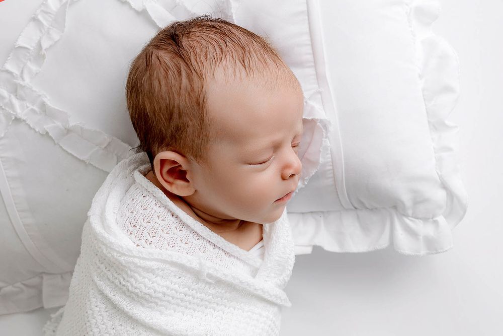 Sleeping newborn baby swaddled in white knit wrap beside ruffled white pillow.