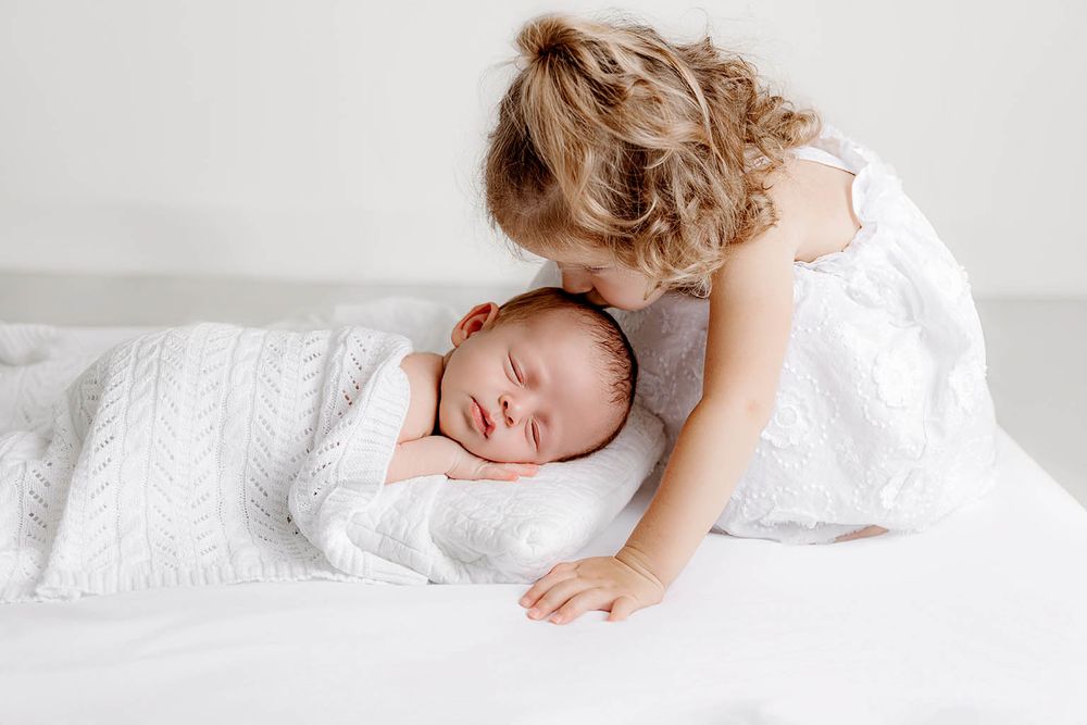 Sweet toddler girl in white dress gently kissing sleeping newborn baby wrapped in white blanket.