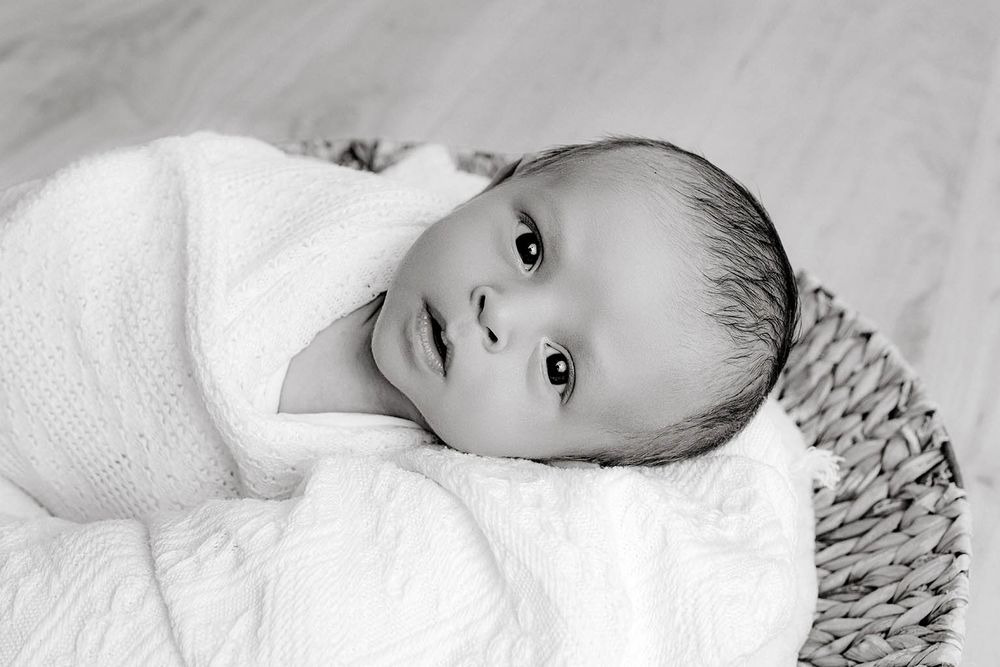 Newborn baby wrapped in white swaddle blanket lying in a wicker basket, black and white photo.