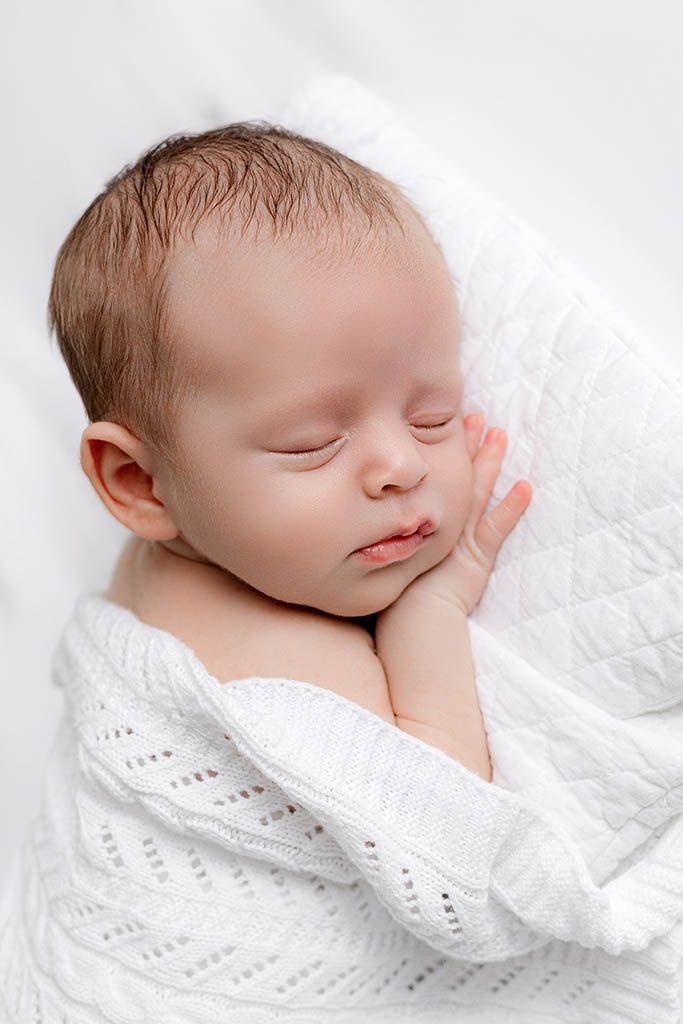 Peaceful newborn baby sleeping wrapped in a soft white knit blanket with tiny hands tucked under cheek.