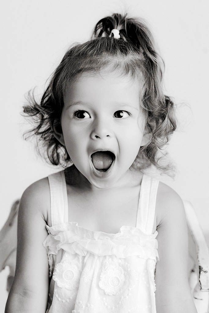 Black and white portrait of surprised toddler girl with curly hair and open mouth wearing white dress.