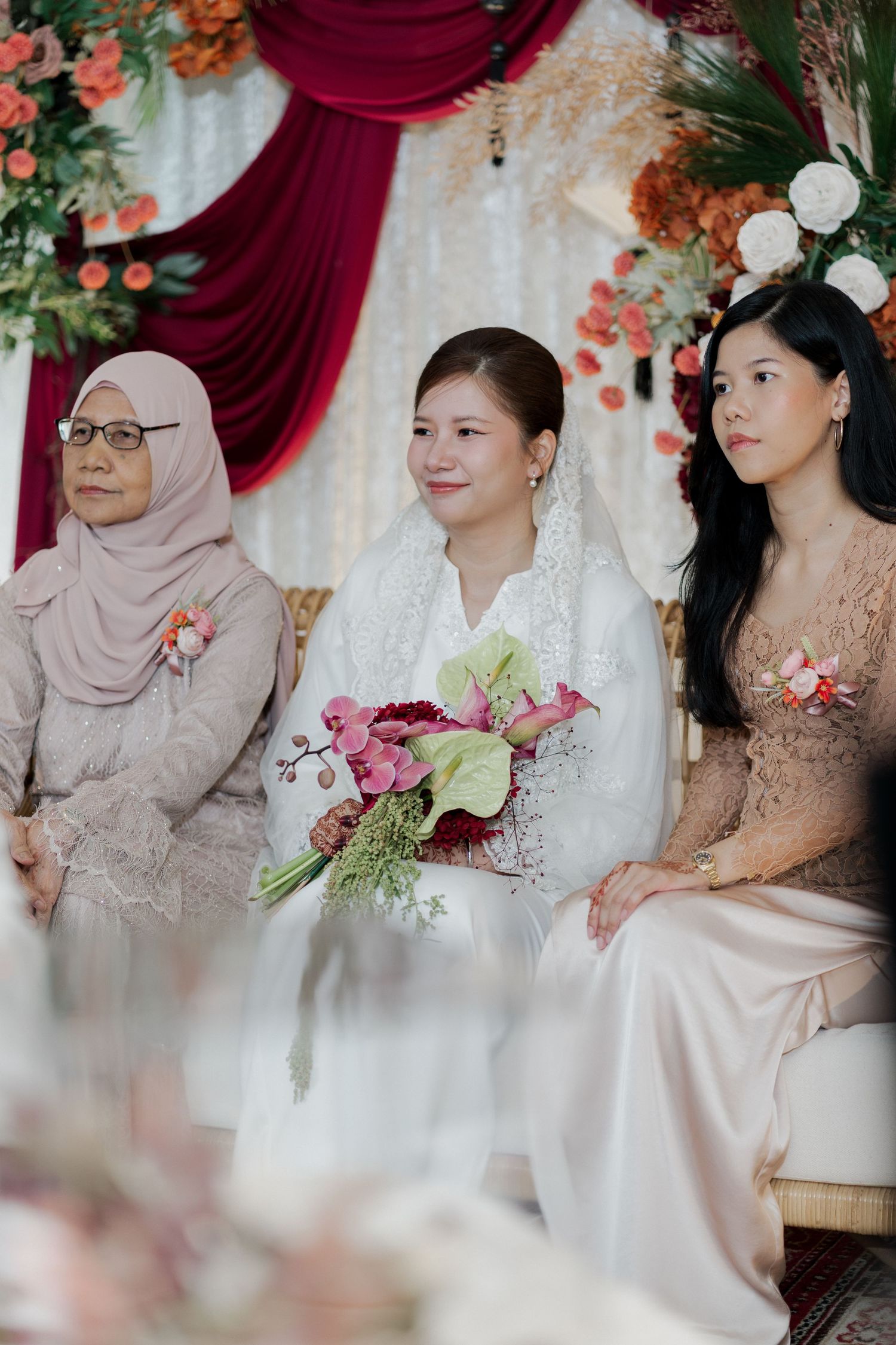 Bride in white lace holding floral bouquet seated between two women in pink at an elegant wedding ceremony.