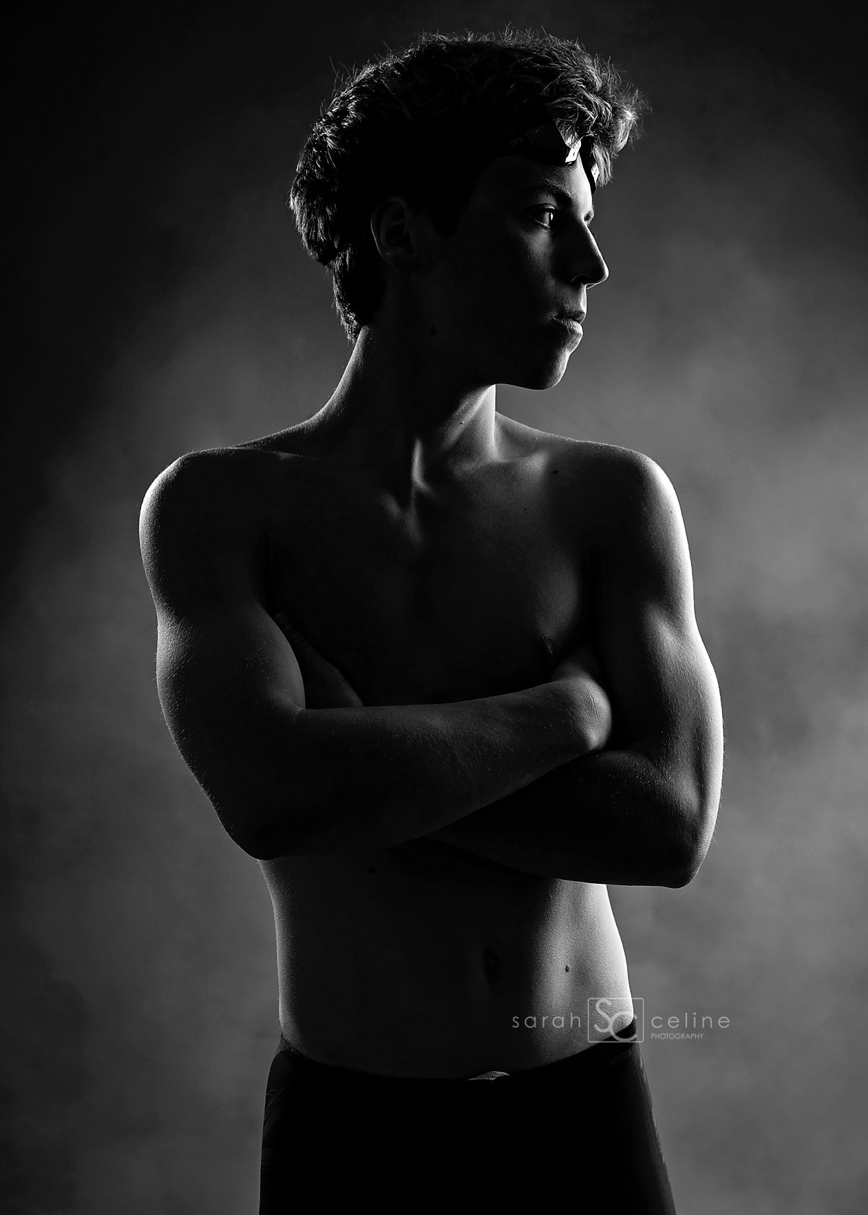 Black and white portrait of a shirtless young man with arms crossed, dramatically lit against a smoky dark background.