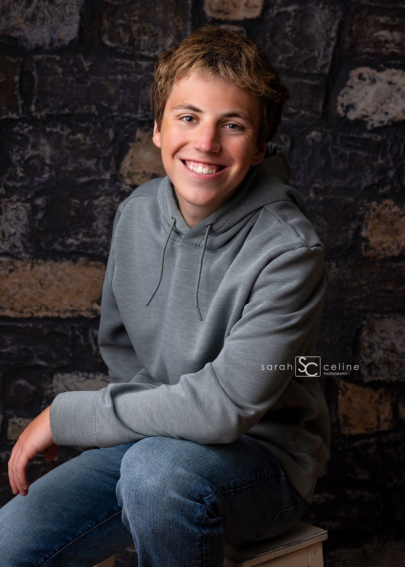 Smiling teenage boy in gray hoodie and jeans poses for senior portrait against rustic stone wall backdrop.