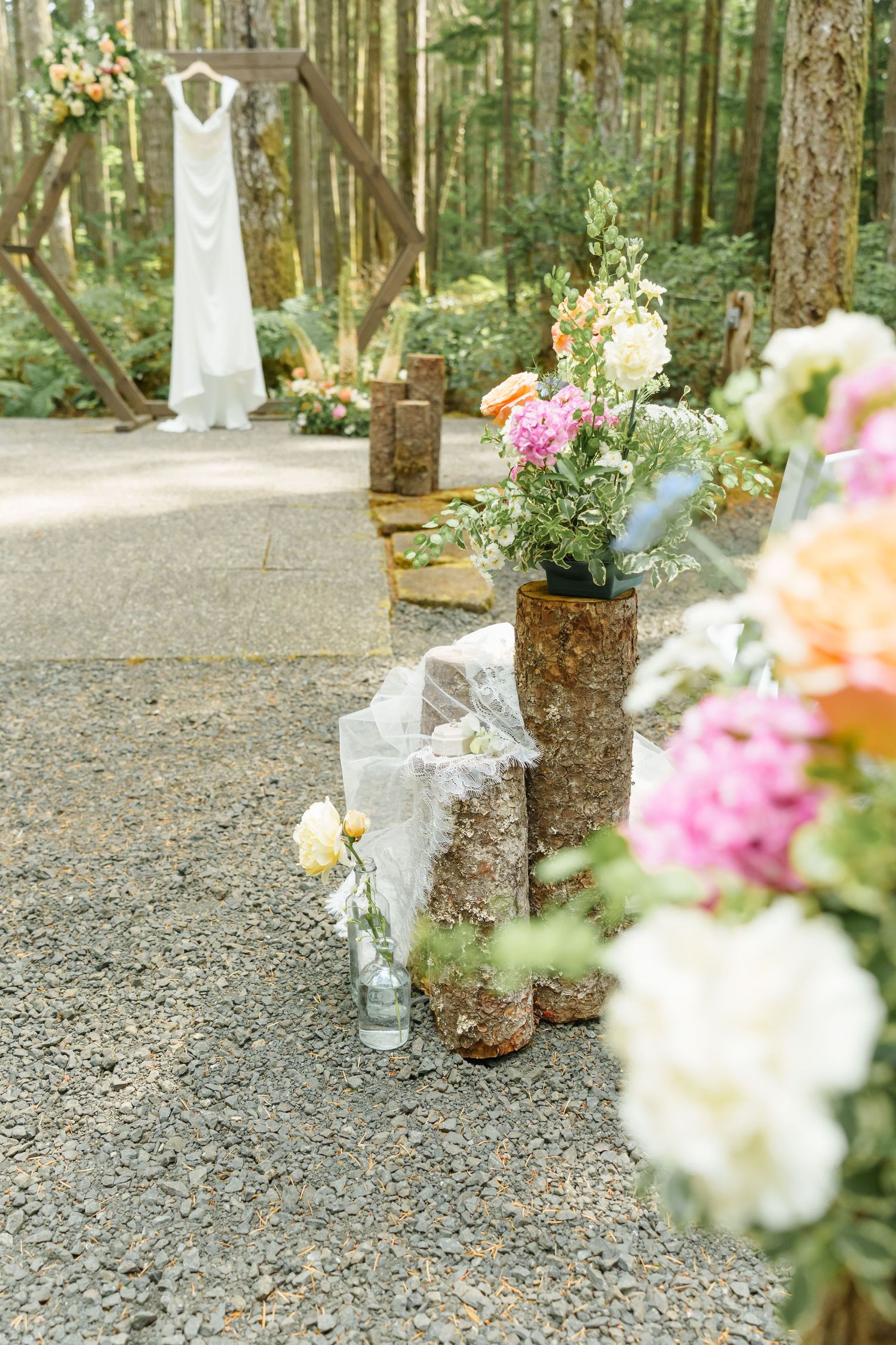 Rustic outdoor wedding aisle with floral arrangements on wood stumps, gravel path, and white gown hanging in forest backdrop.