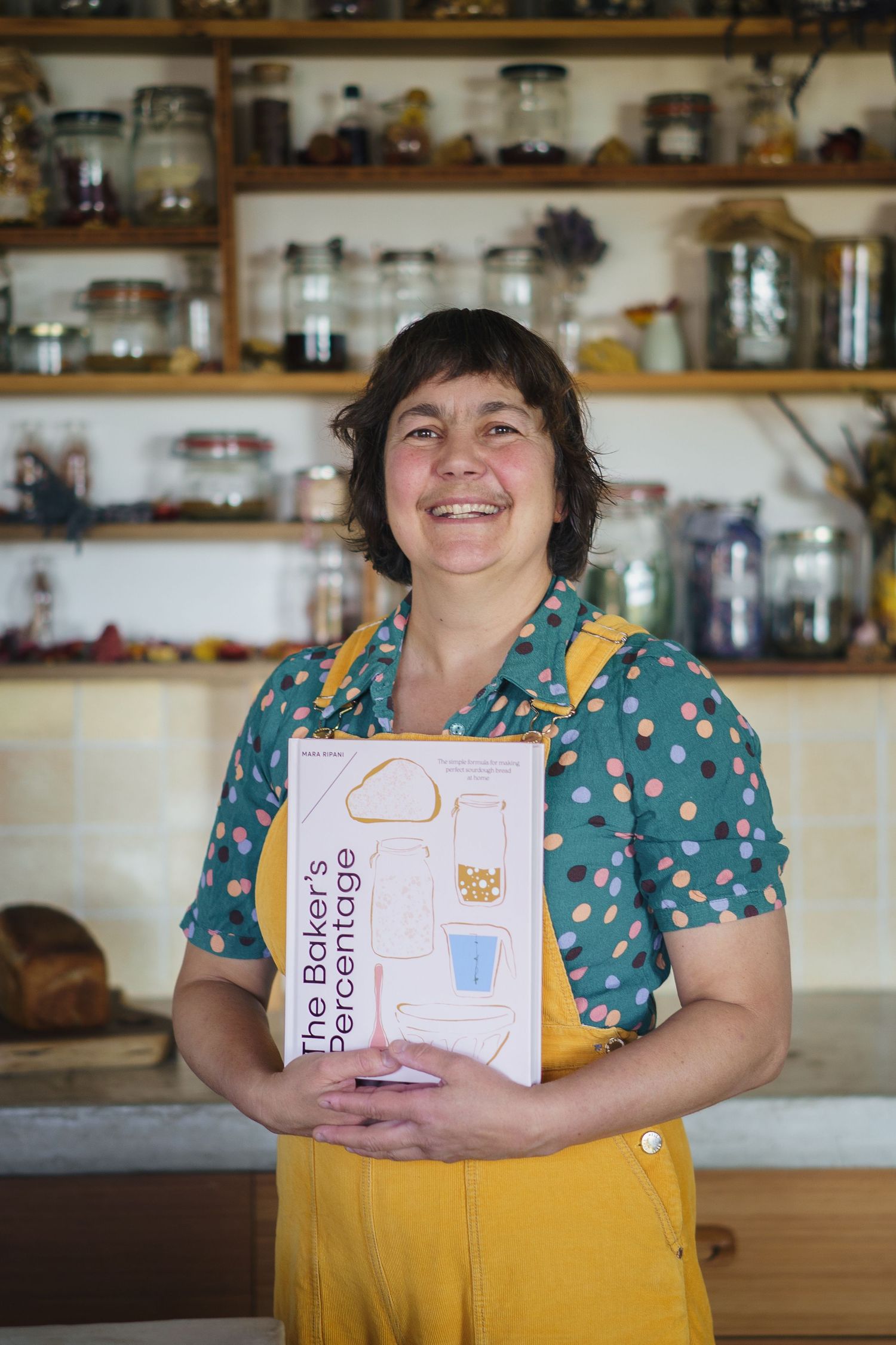 Smiling woman in yellow apron holding The Baker's Percentage cookbook in a rustic kitchen with shelves of jars.

Mara Ripani